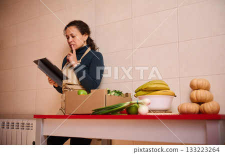 Woman In Apron Reviewing Tablet In A Kitchen With Produce And Pumpkins On Table Woman In Apron Reviewing Tablet In A Kitchen With Produce And Pumpkins On Table 133223264