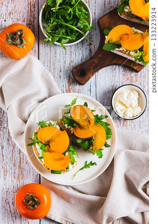 Persimmon, herbs and cream cheese on ciabatta on a plate on the table top and vertical view Persimmon, herbs and cream cheese on ciabatta on a plate on the table top and vertical view 133223914