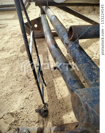 Broken Industrial Ladder on Rusty Pipes. Completely destroyed black metal ladder hanging from heavily rusted pipes in a sandy quarry. Symbol of industrial decay and unsafe conditions. Broken Industrial Ladder on Rusty Pipes. Completely destroyed black metal ladder hanging from heavily rusted pipes in a sandy quarry. Symbol of industrial decay and unsafe conditions. 133224545