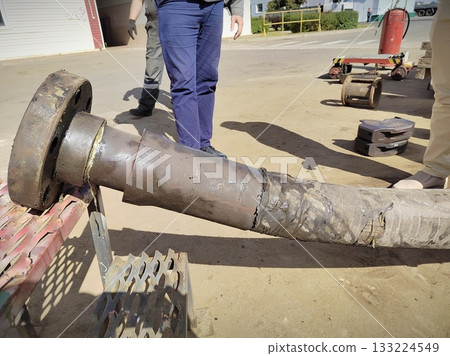 Damaged Heavy Machinery Shaft with Welded Repair. Large worn industrial shaft with visible cracks and rough welding repairs lying on a workbench. Workers in background at outdoor repair yard. Damaged Heavy Machinery Shaft with Welded Repair. Large worn industrial shaft with visible cracks and rough welding repairs lying on a workbench. Workers in background at outdoor repair yard. 133224549