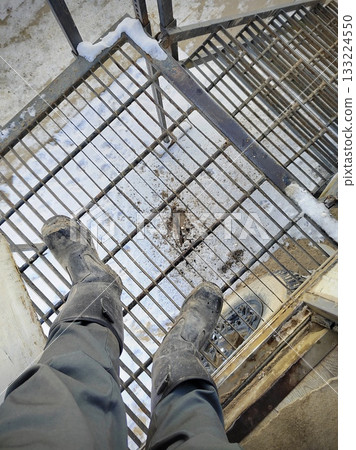 Worker POV on Icy Metal Grating Stairs. First-person view of dirty safety boots on rusty, snow-covered metal grating stairs at an industrial site. Harsh winter conditions, high drop below. 133224550