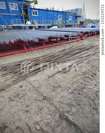 Drilling Rig Pipes and Rebar at Sand Quarry. Stacks of drill pipes on red roller racks and scattered rusty rebar on sandy ground next to a blue industrial cabin with yellow safety railings. 133224552