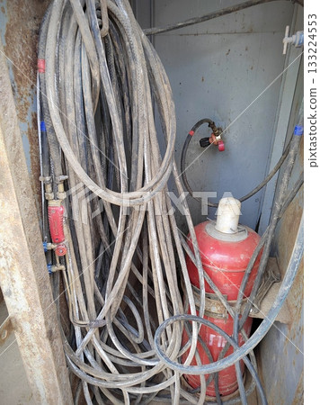 Dirty Welding Hoses and Red Propane Cylinder. Tangled, dust-covered welding-gas hoses hanging next to a worn red propane cylinder with regulator in a grimy industrial compartment. 133224553