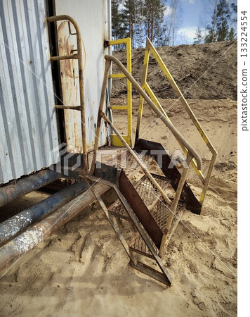 Rusty Metal Stairs to Old Industrial Cabin. Dangerously worn and rusted metal staircase with yellow handrails leading to an old corrugated-metal cabin door in a sandy quarry. 133224554