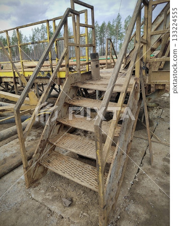 Worn Yellow Metal Stairs on Heavy Machinery Platform. Dirty and rusty yellow metal staircase leading up to an elevated industrial platform surrounded by pipes and forest in the background. 133224555