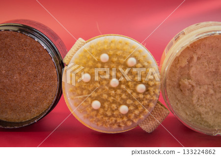 Three jars of body scrub with unique textures and colors, highlighting natural ingredients, arranged on a bright red background, emphasizing skincare and beauty routines 133224862