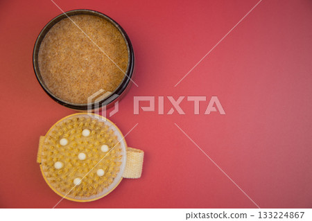Body scrub in black container next to exfoliating brush on pink background, highlighting essential skincare products for a rejuvenating beauty experience 133224867