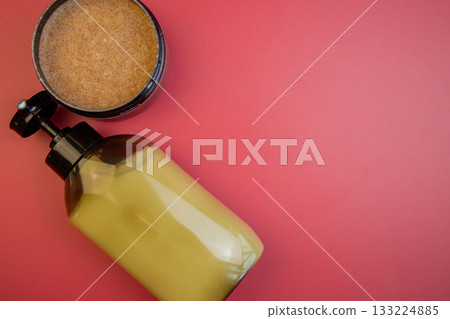 Yellow cosmetic bottle alongside brown sugar scrub in black container on bright red background, highlighting beauty and skincare essentials for personal care 133224885
