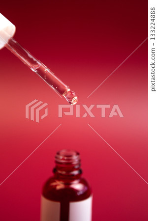 Close-up view of a dropper releasing liquid from a glass bottle, set against a striking red backdrop, highlighting beauty product details and textures 133224888