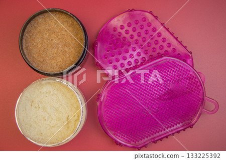 Two body scrub containers, one filled with brown sugar scrub and the other with creamy texture, placed next to a pink exfoliating glove on a colorful background 133225392