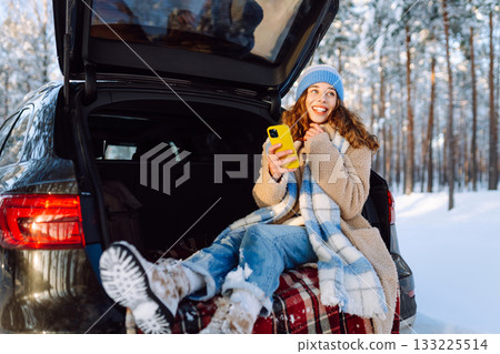 Happy Woman sitting in trunk, using phone. Snowy weather. Concept of adventure, travel by car. Happy Woman sitting in trunk, using phone. Snowy weather. Concept of adventure, travel by car. 133225514