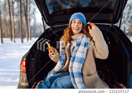 Happy Woman sitting in trunk, using phone. Snowy weather. Concept of adventure, travel by car. 133225517