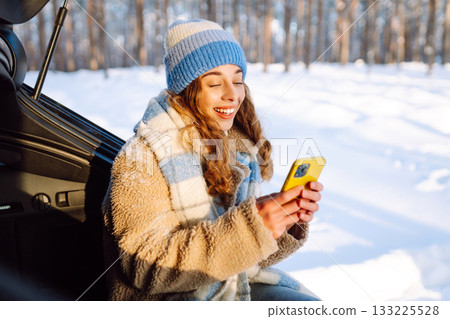 Happy Woman sitting in trunk, using phone. Snowy weather. Concept of adventure, travel by car. 133225528