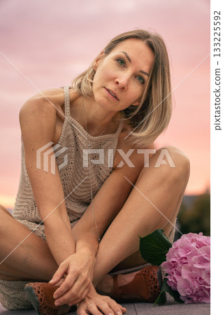 Woman in a beige knitted dress sits thoughtfully on a stone surface at sunset with a pink hydrangea beside her Woman in a beige knitted dress sits thoughtfully on a stone surface at sunset with a pink hydrangea beside her 133225592