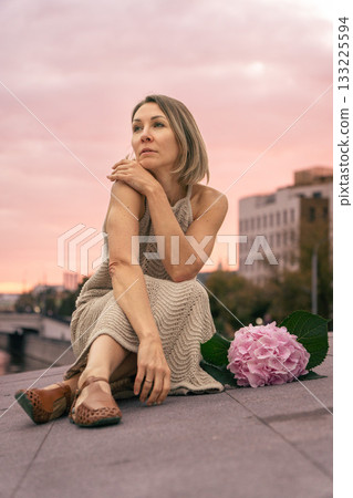 Woman in a beige knitted dress sits thoughtfully on a city embankment at sunset, next to a pink hydrangea. Woman in a beige knitted dress sits thoughtfully on a city embankment at sunset, next to a pink hydrangea. 133225594