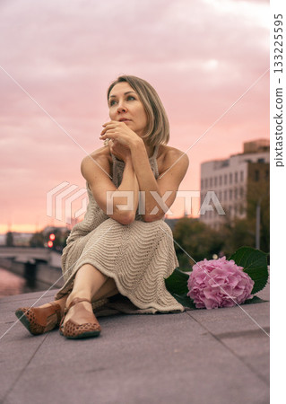 Woman in a beige knitted dress sits thoughtfully on a city embankment at sunset, next to a pink hydrangea. 133225595
