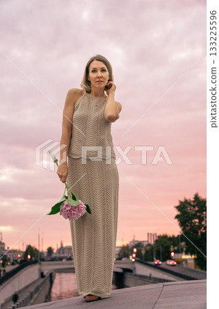 Woman in a knitted beige dress holds a pink hydrangea against a pastel sunset sky over the city 133225596