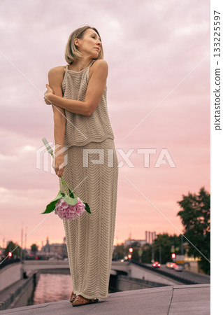 Woman in a knitted beige dress holds a pink hydrangea against a pastel sunset sky over the city Woman in a knitted beige dress holds a pink hydrangea against a pastel sunset sky over the city 133225597