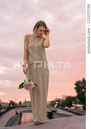 Woman in a knitted beige dress holds a pink hydrangea against a pastel sunset sky over the city Woman in a knitted beige dress holds a pink hydrangea against a pastel sunset sky over the city 133225598