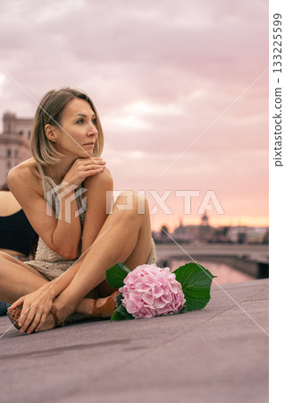 Woman in a beige knitted dress sits thoughtfully on a city embankment at sunset, next to a pink hydrangea. 133225599