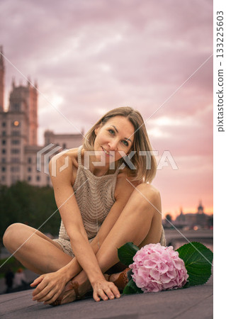 Woman in a beige knitted dress sits thoughtfully on a stone surface at sunset with a pink hydrangea beside her Woman in a beige knitted dress sits thoughtfully on a stone surface at sunset with a pink hydrangea beside her 133225603