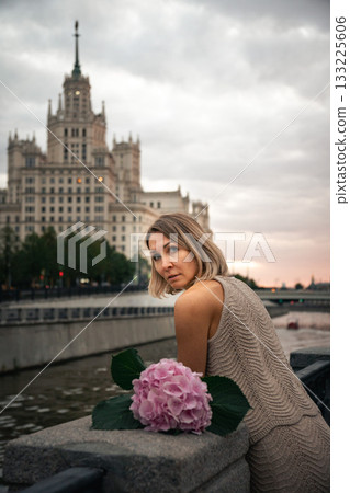 Woman in a knitted beige dress sits by the riverside with a pink hydrangea flower, looking toward a grand city building at sunset 133225606