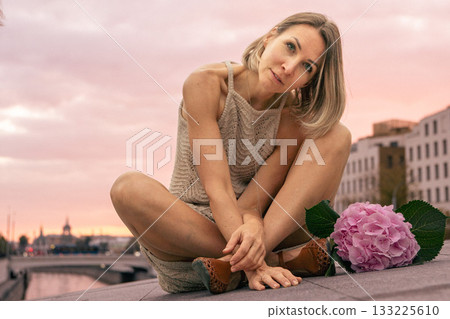 Woman in a beige knitted dress sits thoughtfully on a stone surface at sunset with a pink hydrangea beside her Woman in a beige knitted dress sits thoughtfully on a stone surface at sunset with a pink hydrangea beside her 133225610