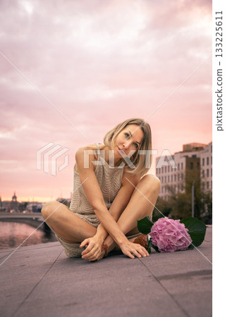 Woman in a beige knitted dress sits thoughtfully on a stone surface at sunset with a pink hydrangea beside her 133225611