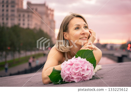 Woman lying on a city embankment at sunset with a pink hydrangea in front of her, smiling softly 133225612
