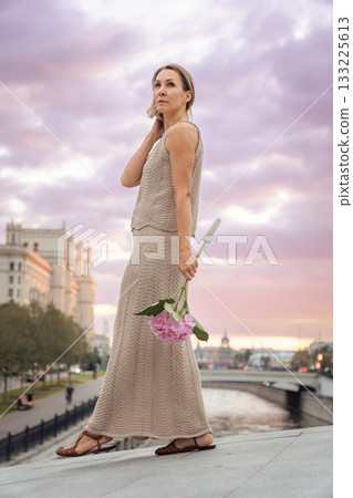 Woman in a beige knitted dress stands near a river holding a pink hydrangea under a dramatic pastel sunset sky Woman in a beige knitted dress stands near a river holding a pink hydrangea under a dramatic pastel sunset sky 133225613