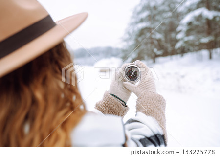 Woman in a hat holding a compass lying in hand on mitten against backdrop of snowy winter forest. Woman in a hat holding a compass lying in hand on mitten against backdrop of snowy winter forest. 133225778