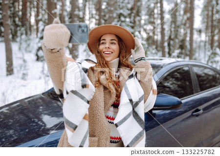 Young tourist takes a selfie with her phone next to her car on a snowy road. Travel concept. Young tourist takes a selfie with her phone next to her car on a snowy road. Travel concept. 133225782