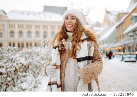 Young happy woman in a winter coat and scarf walks along a snowy city street. Concept of style. Young happy woman in a winter coat and scarf walks along a snowy city street. Concept of style. 133225949