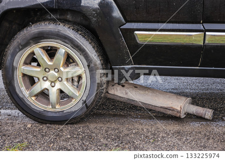 Broken car muffler lying on the road beneath a black vehicle after mechanical damage Broken car muffler lying on the road beneath a black vehicle after mechanical damage 133225974