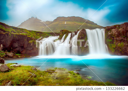 View to the Kirkjufell waterfall at summer. Kirkjufellsfoss waterfall, Iceland, Europe. View to the Kirkjufell waterfall at summer. Kirkjufellsfoss waterfall, Iceland, Europe. 133226662