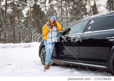 Young woman with phone near her car in a winter forest. Sending messages, communicating with friends 133226669