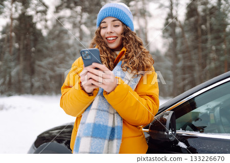 Young woman with phone near her car in a winter forest. Sending messages, communicating with friends Young woman with phone near her car in a winter forest. Sending messages, communicating with friends 133226670