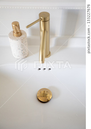 Gold faucet and speckled dispenser on white ceramic sink, polished drain and overflow holes, clean lines and soft reflections convey modern minimal elegance and premium finish 133227676