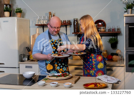 White woman offering taste to man smiling, spoon extended toward partner, playful testing, kitchen counter with bowls, cooperative cooking, candid intimate interaction 133227760