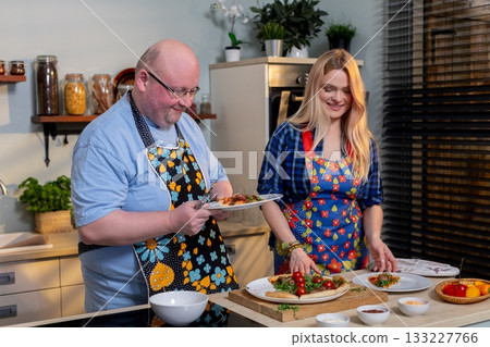 White couple preparing salad together at island, man slicing cucumber, woman arranging greens, colorful vegetables, rustic jars on shelves, upbeat teamwork, healthy 133227766
