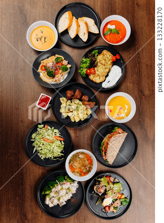 Assorted restaurant dishes flat lay on wooden table with soups salads mains bread and sauces Assorted restaurant dishes flat lay on wooden table with soups salads mains bread and sauces 133228170