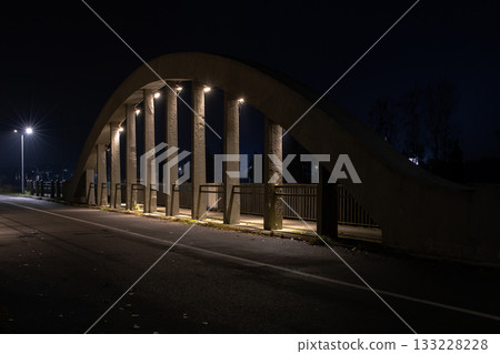 Night view of a concrete arch bridge illuminated by streetlights, with strong shadows and an empty road creating a calm urban atmosphere. Night view of a concrete arch bridge illuminated by streetlights, with strong shadows and an empty road creating a calm urban atmosphere. 133228228