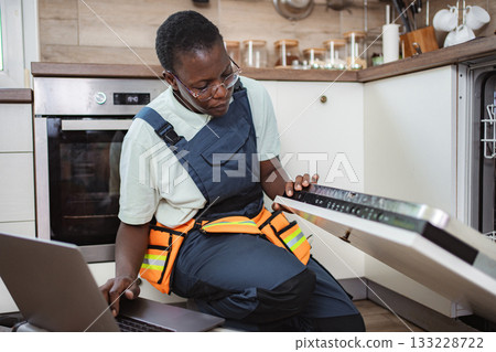 Repairwoman using laptop while repairing dishwasher in kitchen Repairwoman using laptop while repairing dishwasher in kitchen 133228722