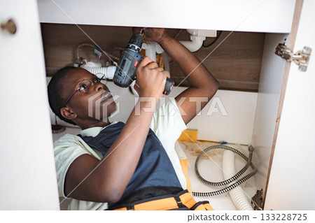Young female plumber lying down and using electric screwdriver beneath kitchen sink 133228725