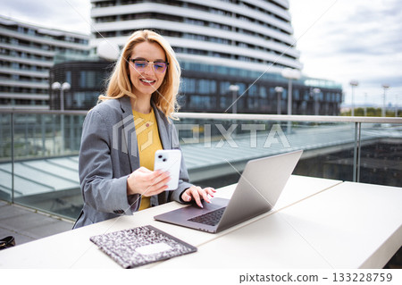 Businesswoman working remotely outdoors, using smartphone and laptop 133228759