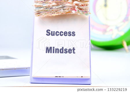 A notepad with the words success mindset stands out with colorful layers. A bright clock in the background adds energy to the motivational setup, encouraging productivity and focus 133229219
