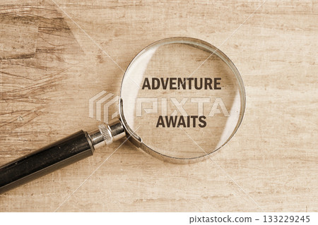 A magnifying glass rests on a rustic wooden surface, focusing on the words adventure awaits. This scene invites curiosity and exploration, sparking the imagination for new journeys ahead 133229245