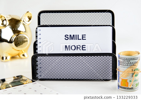 On a clean desk, a note proudly displays the words smile more, inspiring positivity. A piggy bank, a calculator, and rolled cash nearby suggest a focus on saving and financial well-being On a clean desk, a note proudly displays the words smile more, inspiring positivity. A piggy bank, a calculator, and rolled cash nearby suggest a focus on saving and financial well-being 133229333