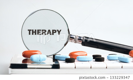 A close-up view shows a magnifying glass focusing on the word therapy, placed on a piano keyboard adorned with colorful pills. This scene creatively links music and mental health support 133229347