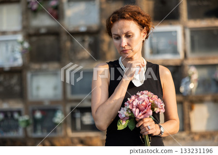 Sad woman in black dress with bouquet of flowers and handkerchief in hands, stands in cemetery 133231196